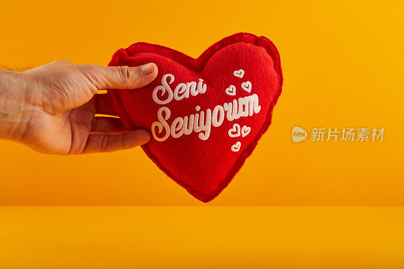 手拿着一个心形的红色填充枕头，上面用土耳其语写着“Seni Seviyorum - I Love You”